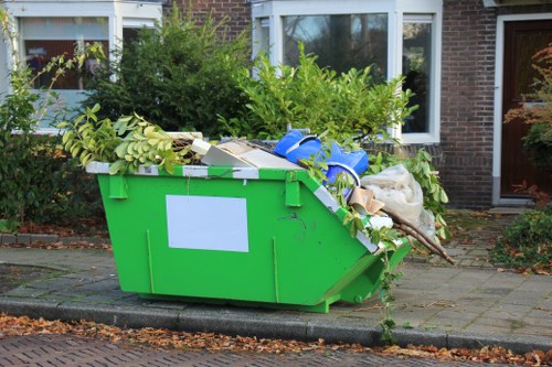 A large bright green plastic skip situated on a paved driveway in front of a residential house with white-framed windows and a brick exterior, filled with a mixture of garden waste including branches, leafy branches, and plant cuttings, alongside some cardboard boxes and white plastic bags. The skip is overflowing with the garden refuse spilling over its edges onto the surrounding pavement, which is lined with a concrete curb. In the background, there are shrubbery and small trees, and the scene is illuminated by natural daylight, indicating a typical outdoor scene in a suburban area suitable for rubbish removal services such as those provided by House Clearance Tring including garden waste clearance and general rubbish disposal.