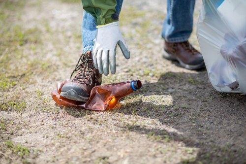 A person wearing white gloves, blue jeans, and brown footwear is seen picking up a used brown glass bottle from a patch of soil or gravel on an outdoor surface. Nearby, there is a large, partially visible plastic rubbish bag on the right side of the image, suggesting a cleanup or rubbish removal activity. The ground appears uneven with a mix of dirt and small stones, indicating a garden, driveway, or outdoor area typical of residential properties. The natural lighting highlights the textures of the soil, the shine on the glass bottle, and the fabric of the person's clothing. This scene reflects a community or professional rubbish clearance activity, aligning with the services offered by House Clearance Tring, a local rubbish removal specialist in the area, focused on maintaining clean and tidy outdoor environments.