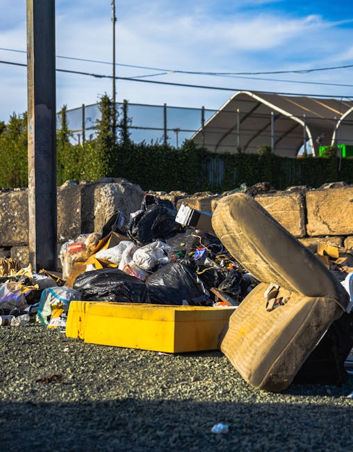 A worker dressed in a green uniform and wearing a safety helmet is standing on the rear platform of a rubbish collection vehicle, which is parked on a residential street. The rear loading compartment 